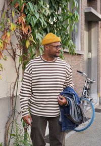 Gestreepte langarmshirt in crème en bruin, gecombineerd met donkerbruine broek. Een gele gebreide muts en een blauwe spijkerjack worden vastgehouden. Groene bladeren op de achtergrond.