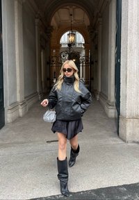 Schwarze Lederjacke mit hohem Kragen, kurzer schwarzer Rock, kniehohe schwarze Stiefel und kleine silberne Handtasche. Dunkle Sonnenbrille getragen.