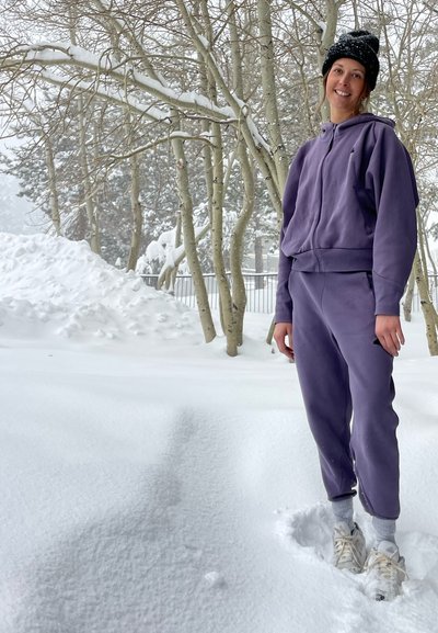 Chándal de forro polar morado con sudadera con cremallera y pantalones ajustados. Lleva puesto un gorro negro, de pie en nieve profunda entre árboles cubiertos de nieve.