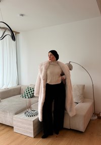 Abrigo peludo beige sobre un suéter de punto crema, combinado con pantalones negros de pierna ancha. El fondo muestra un sofá claro, un cojín verde de cuadros y una lámpara.