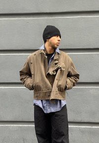 Barna cropped dzseki nagy gombokkal, textúrált gallérral, egy kék kockás ing felett. A szettet fekete sapka és sötét nadrág egészíti ki.