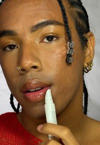 Personne aux cheveux tressés et portant des boucles d'oreilles appliquant un baume à lèvres transparent, vêtue d'un haut rouge sur un fond uni.