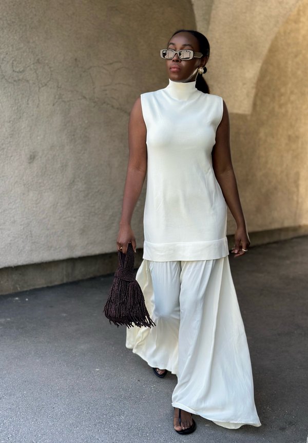 Femme marchant dans une robe blanche sans manches à col montant portée sur un pantalon blanc large, portant de grandes lunettes de soleil et tenant un sac à main tressé foncé.