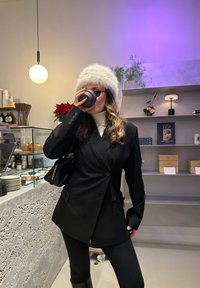 Blazer noir avec de gros boutons, porté sur un col roulé blanc. Chapeau en fausse fourrure grise, sac à main noir, tenant une tasse foncée. Fond de café faiblement éclairé.