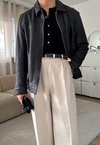 Čierna kožená bunda so zipsom a golierom, spárovaná s čiernou košeľou na gombíky, béžovými nohavicami širokého strihu a čiernym clutchom.