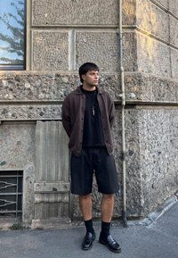 Rudas mygtukinis kardiganas ant juodos marškinėlės, derinamas su juodomis šortais ir loaferiais. Stovi prieš tekstūruotą akmenų sieną.