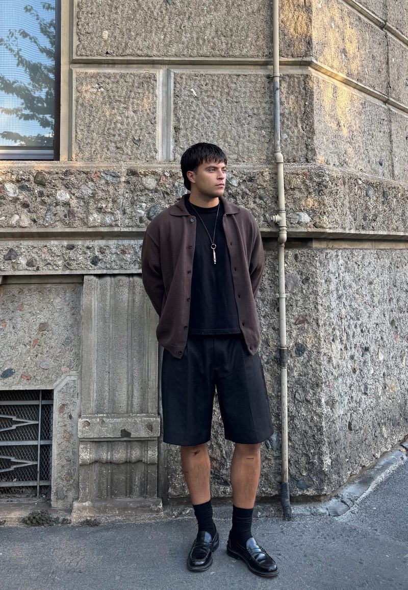 Cardigan marrone con bottoni sopra una maglietta nera, abbinato a pantaloni corti neri e mocassini. In piedi contro un muro di pietra strutturato.