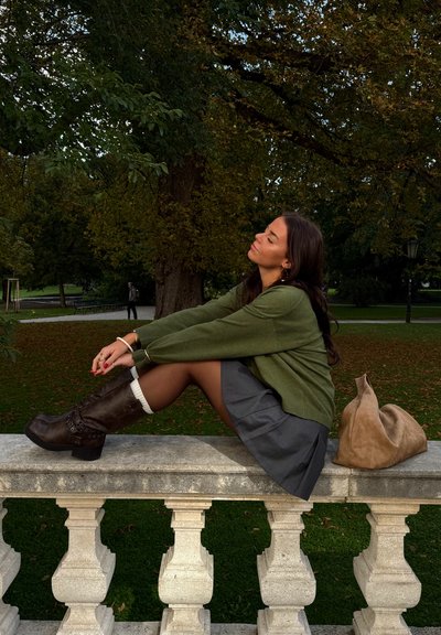 Suéter verde oversize, falda gris, botas marrones hasta la rodilla con adornos, descansando sobre una barandilla de piedra, bolsa beige al lado.
