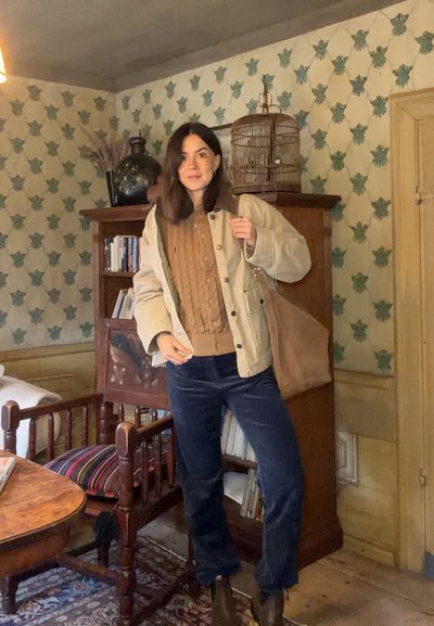 Chaqueta beige con cuello marrón, suéter de punto color beig, pantalones azul marino y bolsa marrón. Muebles de madera y papel tapiz estampado en el fondo.