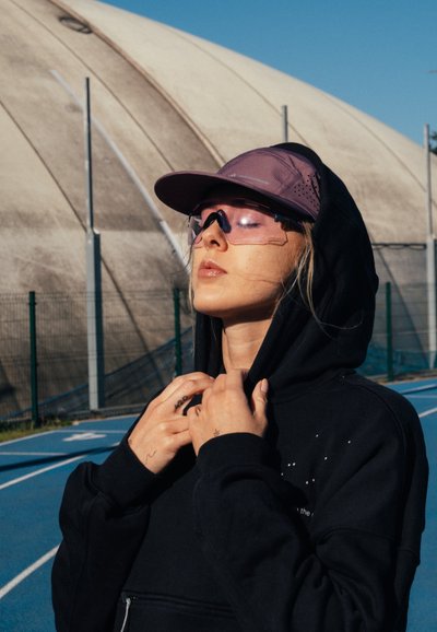 Joven mujer con sudadera negra con capucha y gorra morada ajusta la capucha con los ojos cerrados en una pista de atletismo azul al aire libre bajo un cielo despejado.