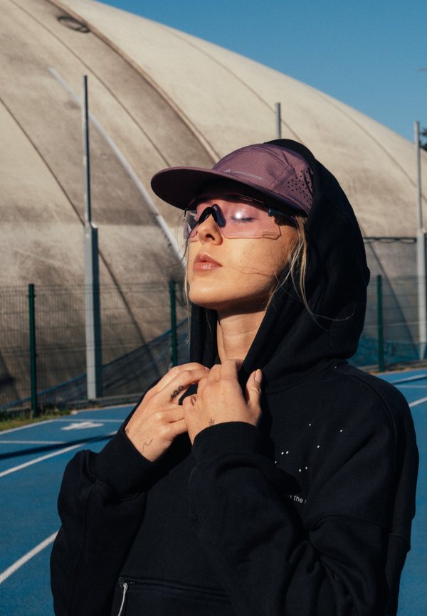 Young woman in a black hoodie and purple cap adjusts her hood with closed eyes on an outdoor blue running track under clear sky.