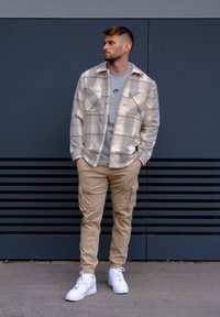 Plastevni outfit, ki vključuje bež karirasto flanelasto srajco, sivo trenirko, bež cargo hlače in bele superge, na sivi podlagi.
