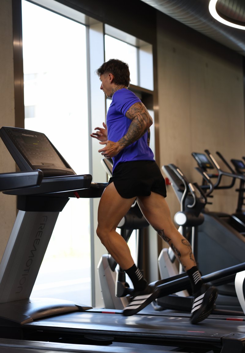 Personne tatouée en chemise bleue et short noir courant sur un tapis de course dans une salle de sport avec des appareils d’entraînement et de grandes fenêtres.