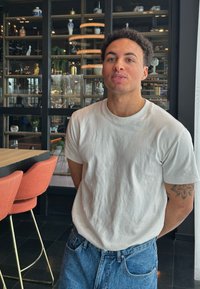 Eine Person trägt ein eng anliegendes weißes Baumwoll-T-Shirt und blaue Jeans und steht in einem modernen Restaurant mit orangefarbenen gepolsterten Stühlen im Hintergrund.