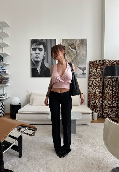 Mujer con camiseta corta rosa y pantalones negros de pie en una sala de estar moderna con sofá blanco, biombo con estampado de leopardo y arte enmarcado en la pared detrás de ella.