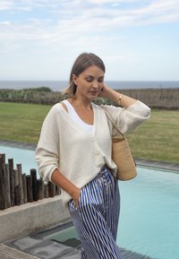 Cardigan lavorato a maglia color crema con scollo a V e bottoni, abbinato a pantaloni a righe blu e bianche. Tiene una borsa di paglia intrecciata.