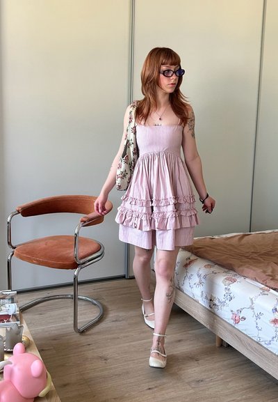 Joven mujer con un vestido rosa con volantes y zapatos blancos caminando en una habitación con una silla y una colcha floral.