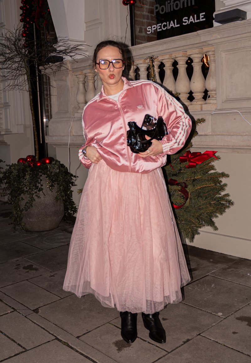 Veste bomber satinée rose avec rayures blanches, jupe en tulle superposée, bottes pointues noires, et tenant une pochette noire. Décorations de fêtes en arrière-plan.