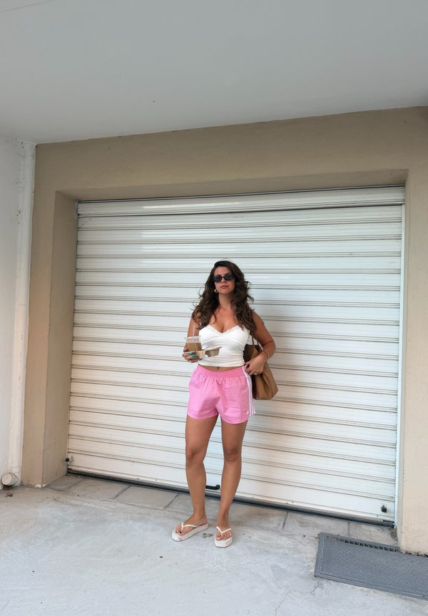 Femme portant des lunettes de soleil, un haut blanc et un short rose, debout tenant une tasse de café et un sac marron devant une porte de garage blanche fermée.