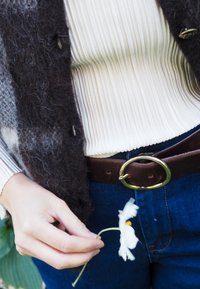 Gilet grigio scuro alla moda con un top crema a coste sotto, cintura in pelle marrone con una fibbia circolare dorata, che tiene un fiore bianco.