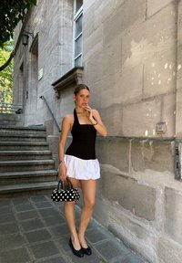 Top negro de cuello halter con una falda blanca con volantes, acompañado de un bolso de lunares y zapatos negros. El fondo incluye una pared de piedra y escaleras.