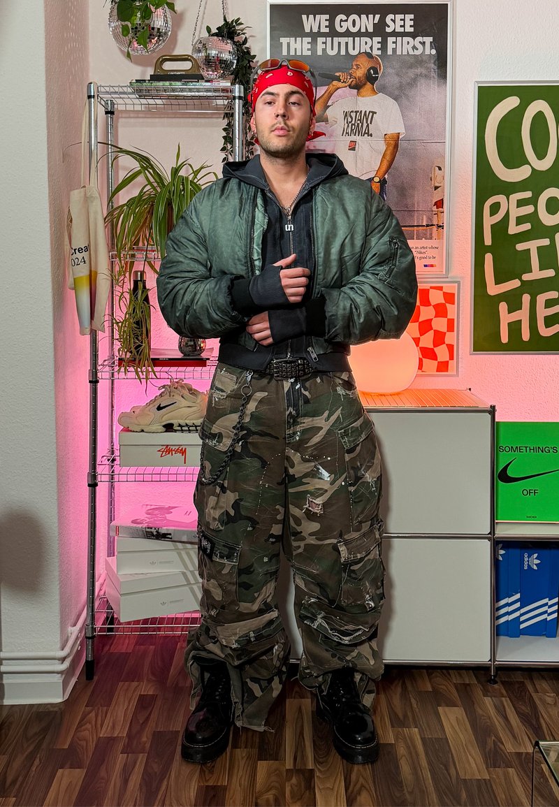 Bomber jacket in dark green, layered over a black top; camo cargo pants with pockets and chains; black chunky boots; red bandana.