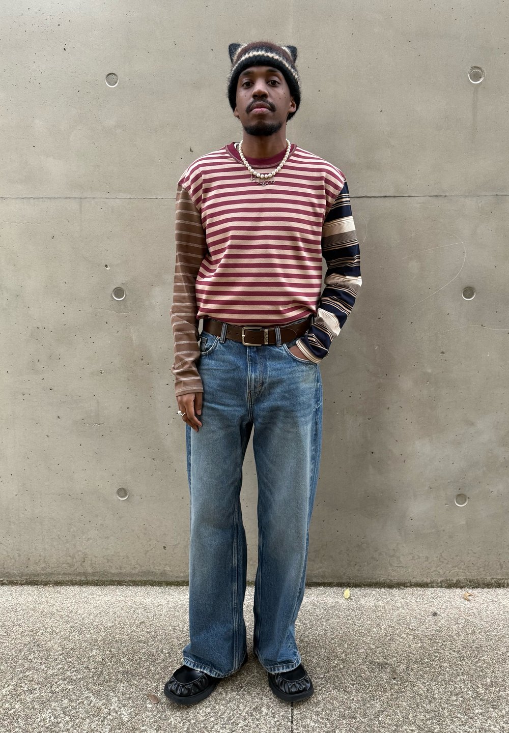 Koszula w paski z długim rękawem w kolorach bordowym i brązowym, zestawiona z jasnoniebieskimi szerokimi dżinsami oraz czarnymi butami wsuwanymi. Do tego ciemna czapka beanie.