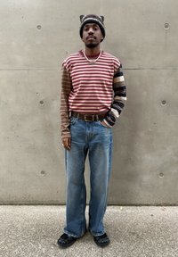 Koszula w paski z długim rękawem w kolorach bordowym i brązowym, zestawiona z jasnoniebieskimi szerokimi dżinsami oraz czarnymi butami wsuwanymi. Do tego ciemna czapka beanie.