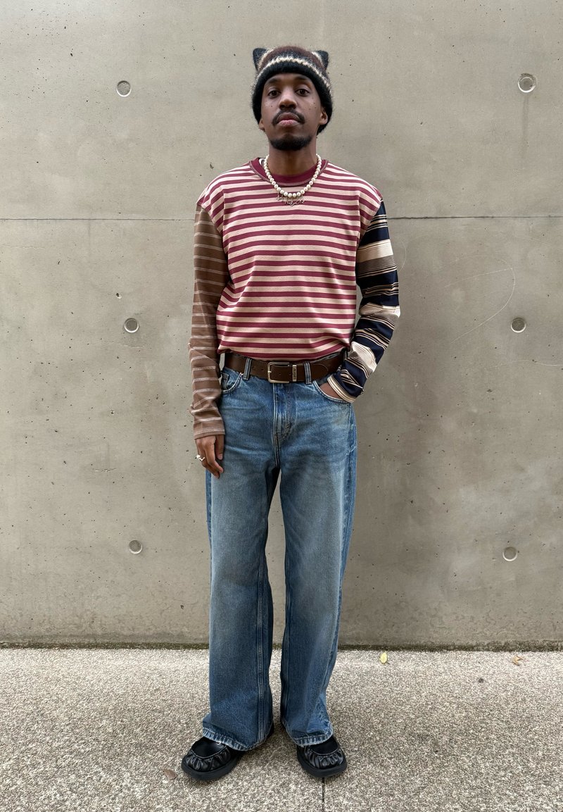 Koszula w paski z długim rękawem w kolorach bordowym i brązowym, zestawiona z jasnoniebieskimi szerokimi dżinsami oraz czarnymi butami wsuwanymi. Do tego ciemna czapka beanie.