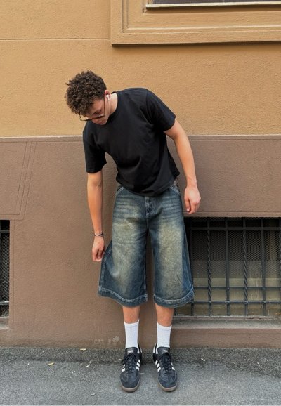 Camiseta negra, pantalones cortos de denim azul claro, calcetines blancos y zapatillas negras. La persona está de pie en una acera cerca de una pared marrón texturizada.