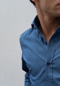 Modra denim srajca s ostrim ovratnikom, dolgimi rokavi in enojnim gumbom za zapiranje. Opazna podrobnost: majhna luknja blizu drugega gumba.
