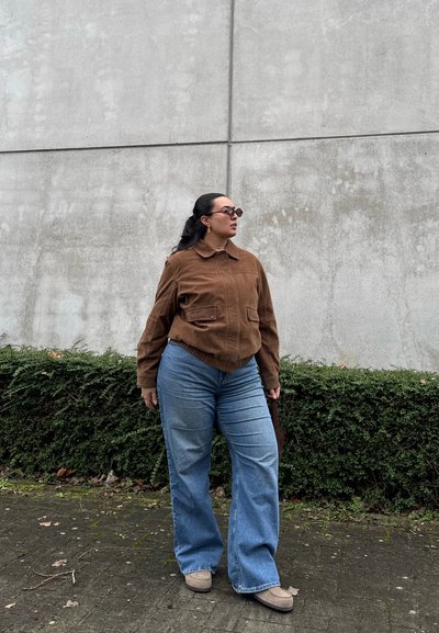 Mujer con cabello oscuro recogido en una cola de caballo, usando gafas de sol, una chaqueta marrón, jeans azules y zapatos beige, de pie en una acera cerca de arbustos verdes.