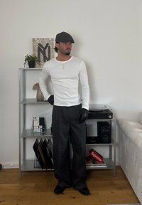 Férfi fehér hosszú ujjú ingben, fekete kesztyűben, nadrágban és sapkában áll egy fém polc mellett, amelyen növények, lemezek és audio berendezések találhatók egy nappaliban.