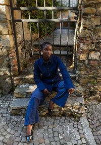 Completo in denim con una camicia e pantaloni a gamba larga di colore blu scuro, dotato di colletto e bottoni. Tacchi a punta neri, seduta su gradini di pietra.