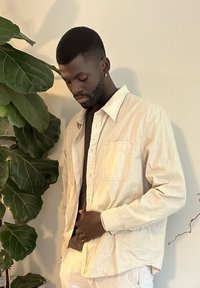 Camicia button-up beige chiaro sopra un maglione a maglia scuro, con taschino sul petto e maniche arrotolate, accanto a una grande pianta con foglie.