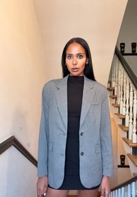 Blazer oversized cinza em padrão xadrez com ombros estruturados, duas bolsos frontais, fecho de botão único, usado sobre um vestido preto. Textura de tecido suave.