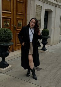 Czarny dwurzędowy płaszcz na białej koszuli, w połączeniu z beżową spódnicą i czarnymi botkami z wydłużonym czubkiem. Stoi w pobliżu drewnianych drzwi i rośliny.