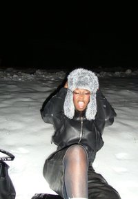 Personne portant une tenue en cuir noir et un chapeau en fourrure grise assise sur la neige la nuit, tenant le chapeau des deux mains et la bouche ouverte.