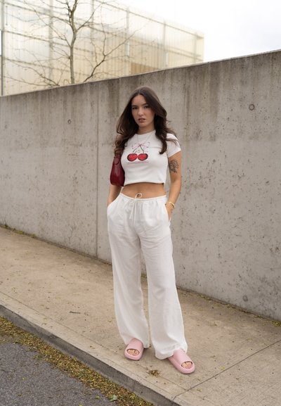 Camiseta blanca cortada con gráfico de cereza, pantalones amplios color crema, sandalias rosas y un bolso rojo. El fondo presenta una pared de concreto.