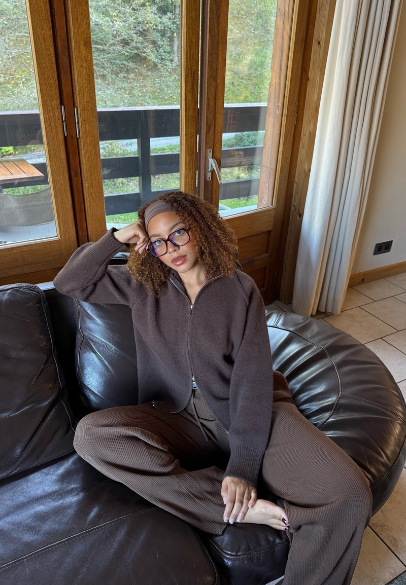 Maglione marrone con zip sopra pantaloni a coste abbinati, seduto su un divano di pelle con le mani posate sulle ginocchia. Finestre in legno e illuminazione soffusa.