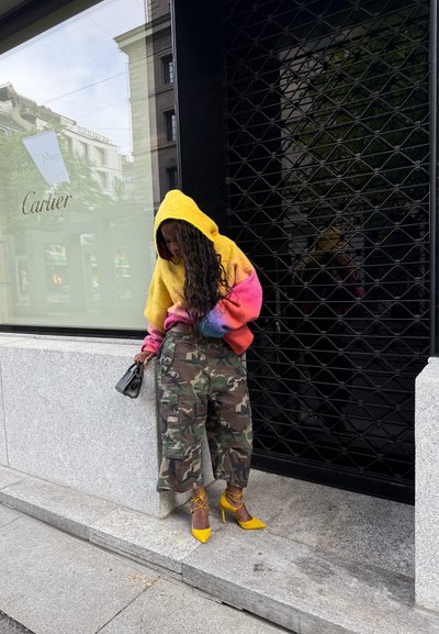 Un suéter con capucha grande y colorido que hace una transición de amarillo a rosa, combinado con pantalones cargo de camuflaje y zapatos de tacón alto de un amarillo brillante.