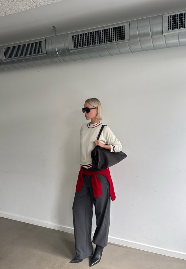Une femme portant des lunettes de soleil, un pull blanc, un pantalon gris et des bottes noires porte un sac à main noir dans un espace intérieur sobre.