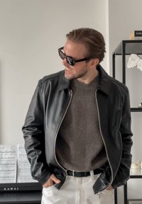 Czarna skórzana kurtka na zamek, szary sweter z prążkowanym wzorem oraz beżowe spodnie. Dodatki to ciemne okulary przeciwsłoneczne i czarny pasek.