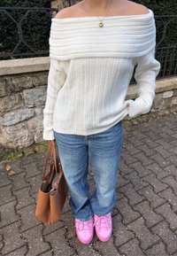 Balts ribots piegriezums džemperis ar garajiem piedurknēm, gaiši zilas džinsu bikses, rozā kurpes un brūna soma uz akmeņaina fona.