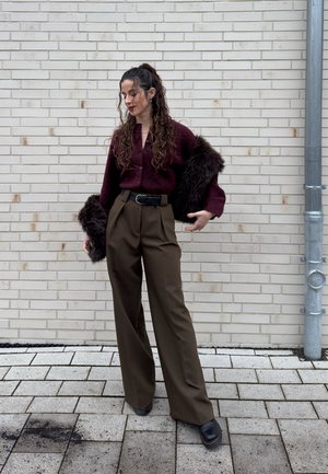 Mujer con cabello largo y rizado, que lleva un suéter burdeos, pantalones anchos marrones y botas negras, de pie contra una pared de ladrillo claro.