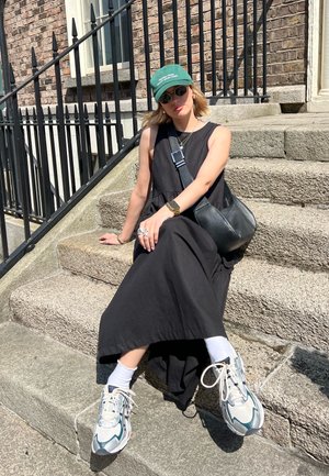 Vestido negro sin mangas, zapatillas gruesas blancas y grises, gorra verde. Entorno casual en escalones de piedra, con un bolso negro cruzado.
