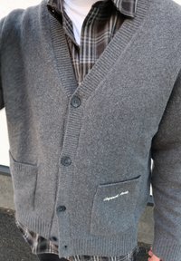 Cardigan gris en tricot avec un col en V, deux poches avant et trois boutons. Superposé à une chemise à carreaux avec un col rond blanc en dessous.