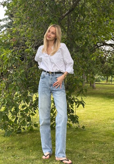 Camisa blanca de botones, jeans de tiro alto de color azul claro y cinturón negro. De pie en un entorno exterior verde con árboles y hierba.