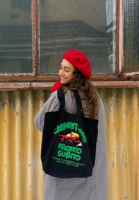 Femme portant un béret rouge et un manteau gris sourit de côté, tenant un sac cabas noir avec un texte graphique coloré et une illustration.