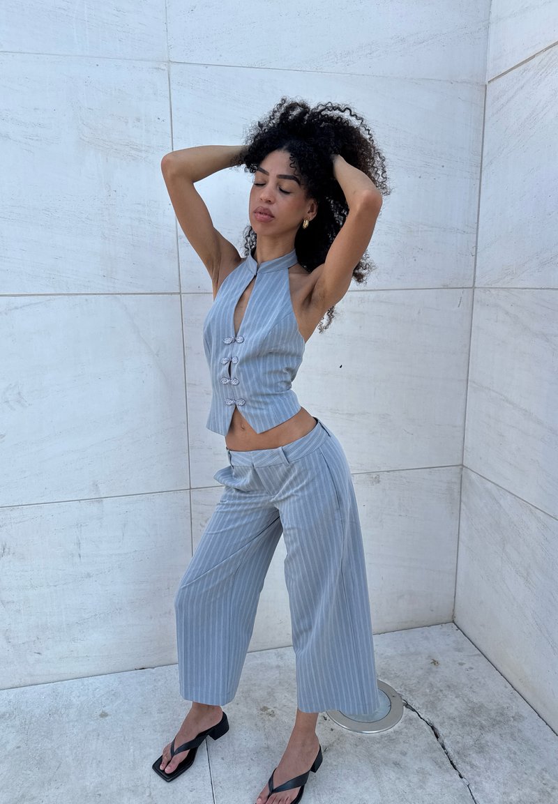 Femme aux cheveux bouclés portant un gilet sans manches à fines rayures bleu clair et un pantalon large assorti, posant avec les mains dans les cheveux contre un mur de pierre clair.